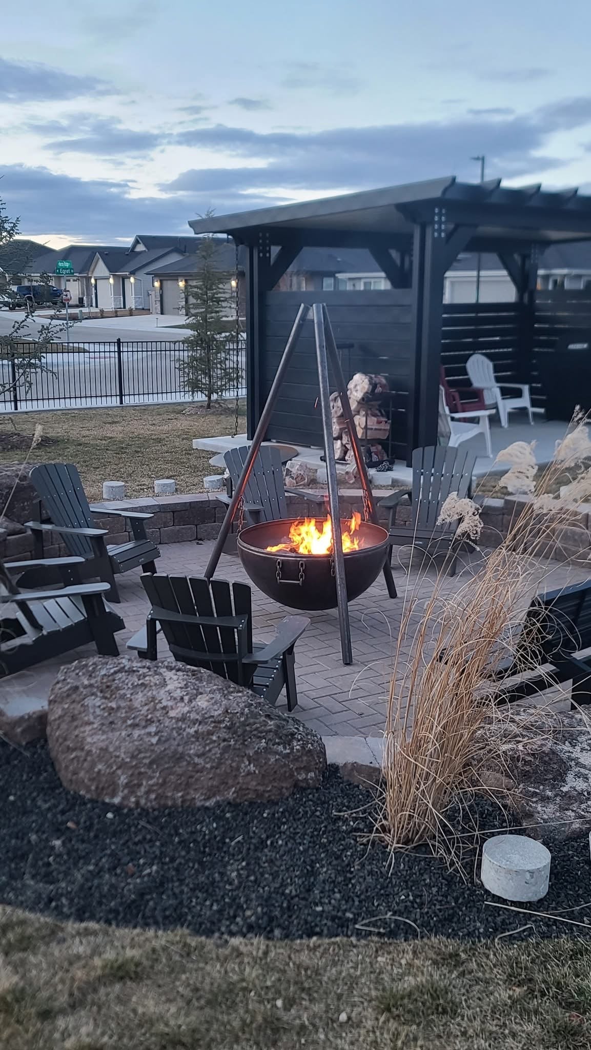 A backyard fire pit area at dusk featuring a circular stone patio with several Adirondack chairs arranged around a modern tripod fire pit. The fire pit emits a warm, inviting glow. There is greenery, tall trees, and a house with glowing windows in the background, creating a relaxing outdoor atmosphere.