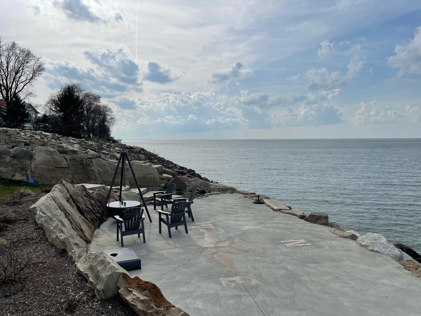 A cozy firepit scene by a tranquil lake at dusk. The stone firepit is surrounded by rustic wooden Adirondack chairs; soft, golden flames flicker and reflect on the gentle lake waters. Tall pine trees frame the background, while fireflies dance in the air. There are marshmallows on sticks ready for roasting, and a plaid blanket draped over one chair. The sky glows in warm tones of orange and purple as the sun sets over the distant hills. The mood is peaceful, inviting, and perfect for a summer evening by the